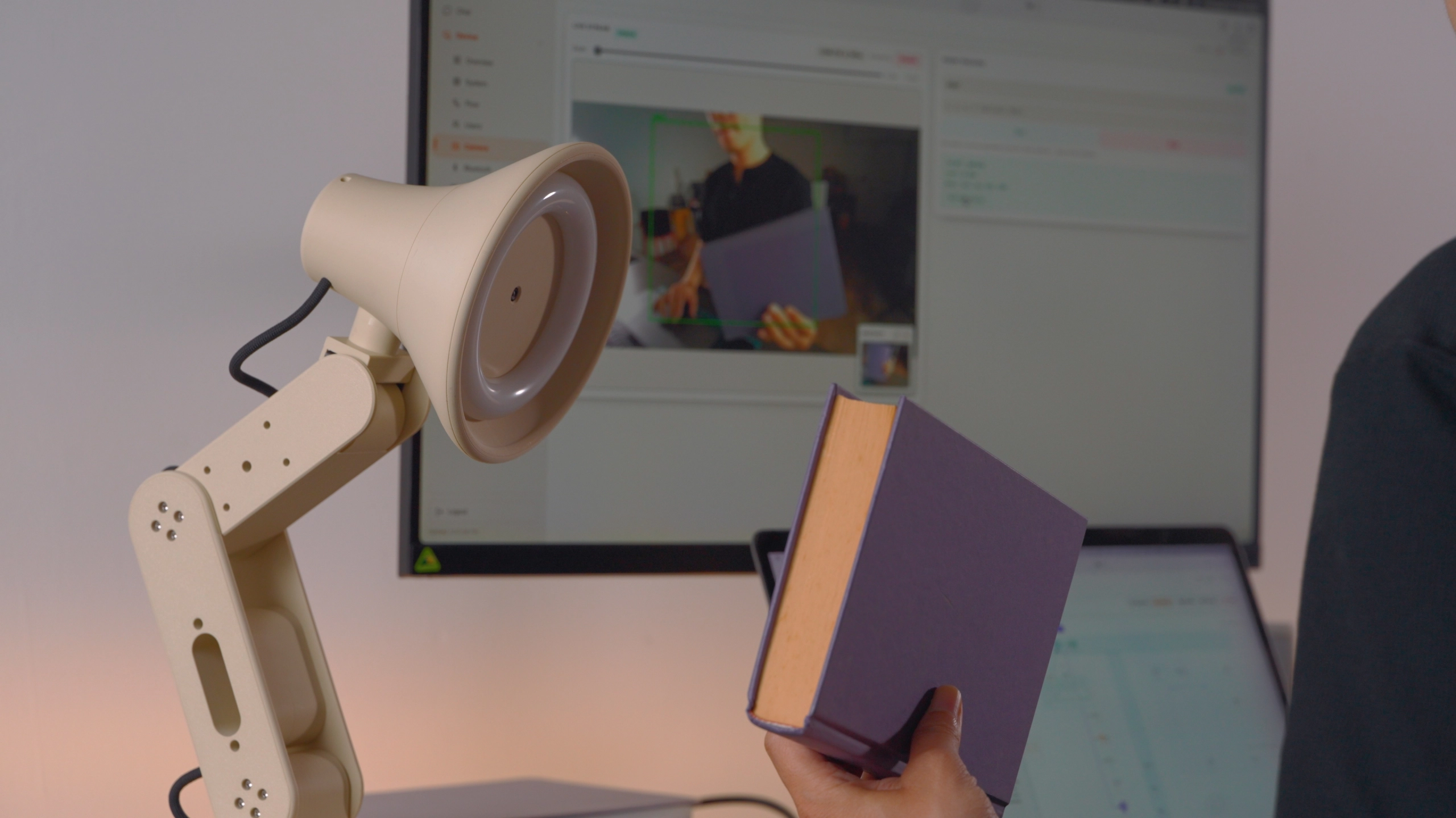 A person holds up a book for Lamp, while a screen behind shows what Lamp’s camera sees