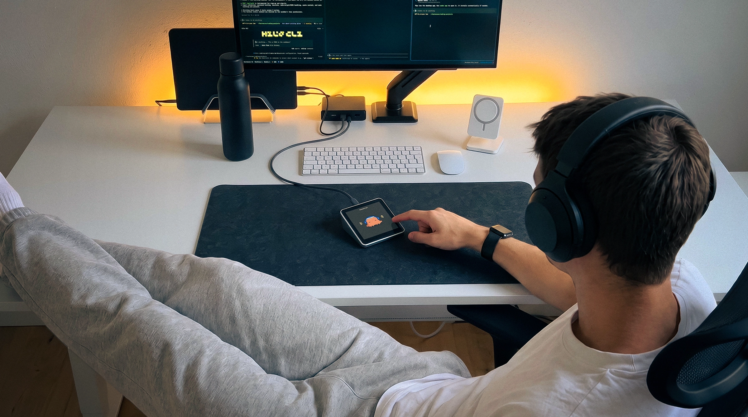 Someone leaning back at a white desk with headphones on, tapping Harness Pro on the desk mat, a monitor full of agents behind it.